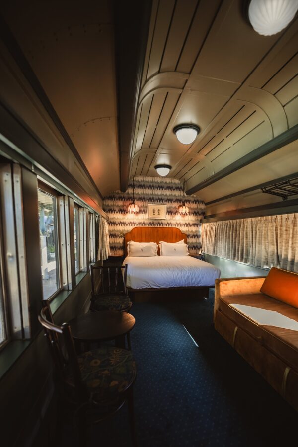 5 Star Hotels in Chattanooga, Tennessee to Book Now. 5 Interior of a restored vintage train car suite with a cozy double bed, retro lighting, patterned wallpaper, and a velvet orange loveseat. Large windows line one side, letting in natural light. A small round table with two floral-upholstered chairs sits near the window, creating a charming and nostalgic atmosphere.