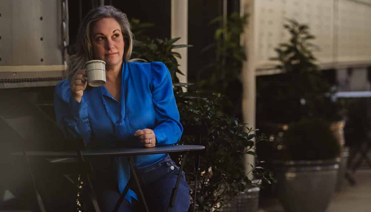 Kathy sipping coffee at a vintage table between two restored sleeper train cars at Hotel Chalet in Chattanooga. She’s wearing a blue top and looks relaxed, enjoying a peaceful morning moment during her stay.