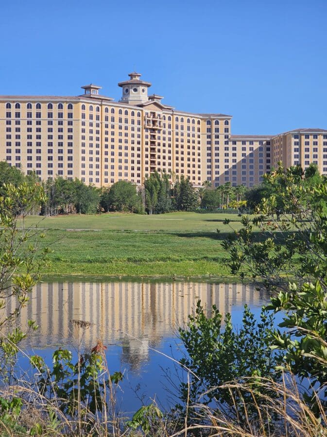 Distant view of Rosen Shingle Creek resort in Orlando, Florida, reflected in a calm pond with greenery in the foreground and a bright blue sky above.