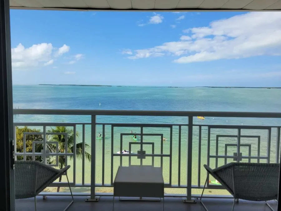 Baker's Cay Resort Key Largo review 4 Balcony view from a Baker’s Cay Resort room in Key Largo, Florida, overlooking turquoise waters, paddleboarders, and a clear blue sky with two chairs and a small table in the foreground