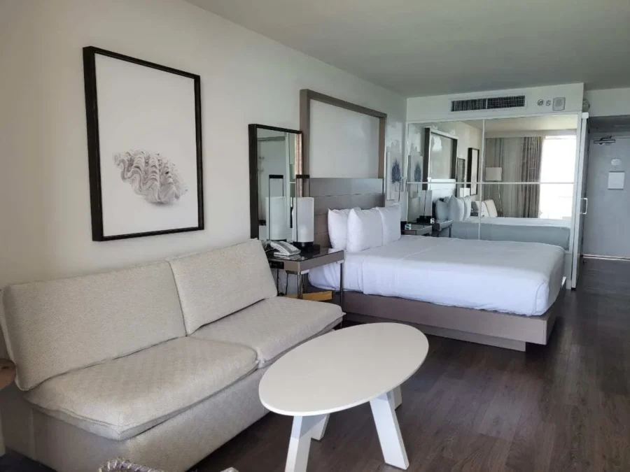 Baker's Cay Resort Key Largo review 3 Modern guest room at Baker’s Cay Resort in Key Largo featuring a king bed, a light gray sofa, a round white coffee table, wood floors, and coastal artwork with a mirrored wall.