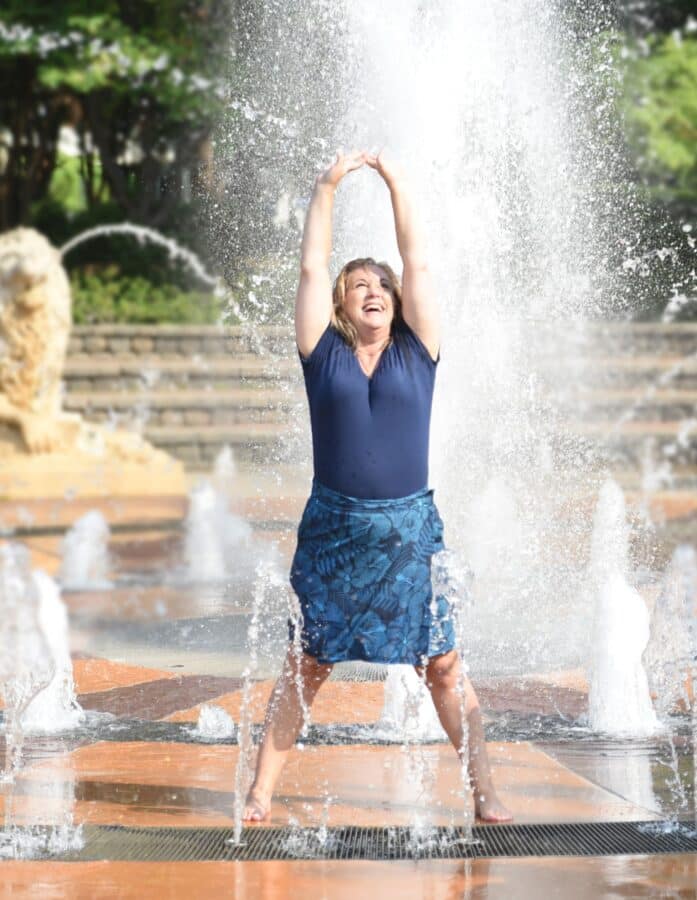 Where to Go on a Weekend Trip: 50 Easy Getaways in the USA 11 Woman standing barefoot in a splash fountain with arms raised, smiling joyfully. Sheβs wearing a navy blue top and floral skirt, surrounded by streams of water shooting up from the ground on a sunny day. Trees and a stone lion fountain are visible in the background.