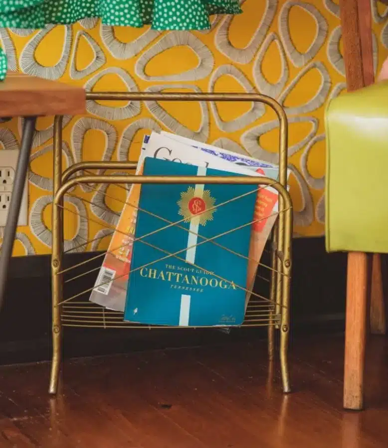 Quirky, Cool, and Totally Chattanooga: Inside The Dwell Hotel (Review) 2 A vintage-style magazine rack at The Dwell Hotel in Chattanooga, TN holding a copy of The Scout Guide Chattanooga, set against retro yellow wallpaper and hardwood floors.