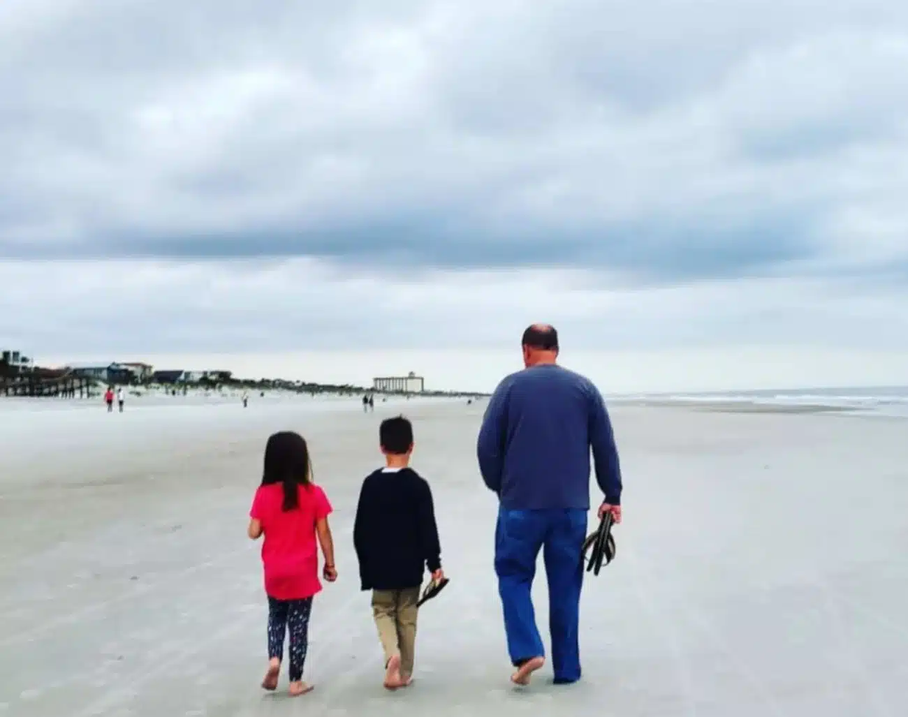 A man walking barefoot with two children along the wide, uncrowded shoreline of Neptune Beach, Florida under a cloudy sky.