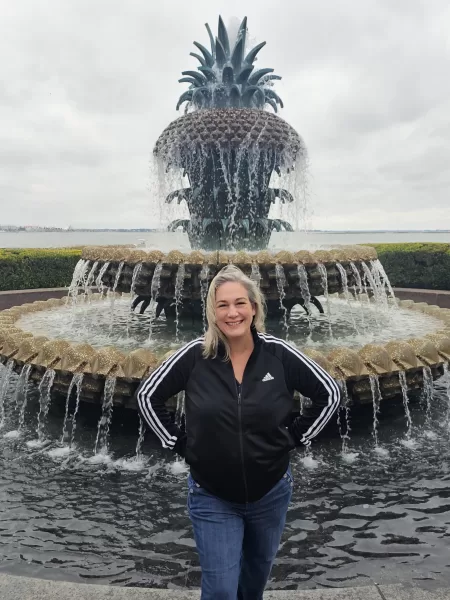 The Ultimate Charleston Weekend Getaway for Women: Walkable, Charming & Full of Southern Surprises 9 Travel blogger Kathy Brown stands smiling in front of the iconic Pineapple Fountain in Charleston, South Carolina, wearing a black Adidas jacket and jeans on a cloudy day.