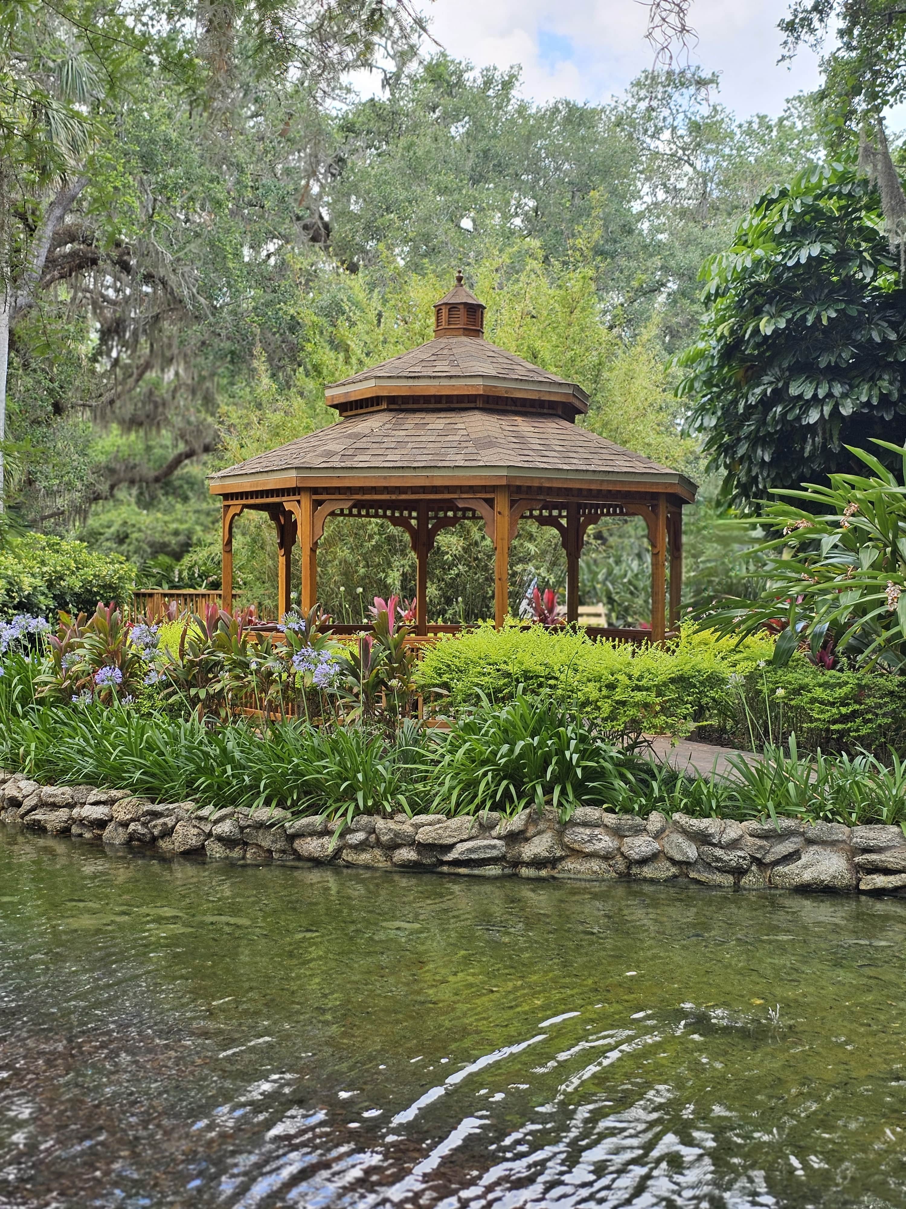 A charming wooden gazebo surrounded by lush tropical plants and colorful flowers sits beside a gently flowing stream at Washington Oaks Gardens State Park in Florida. Tall oak trees and vibrant greenery create a peaceful, shaded retreat in this scenic garden setting.