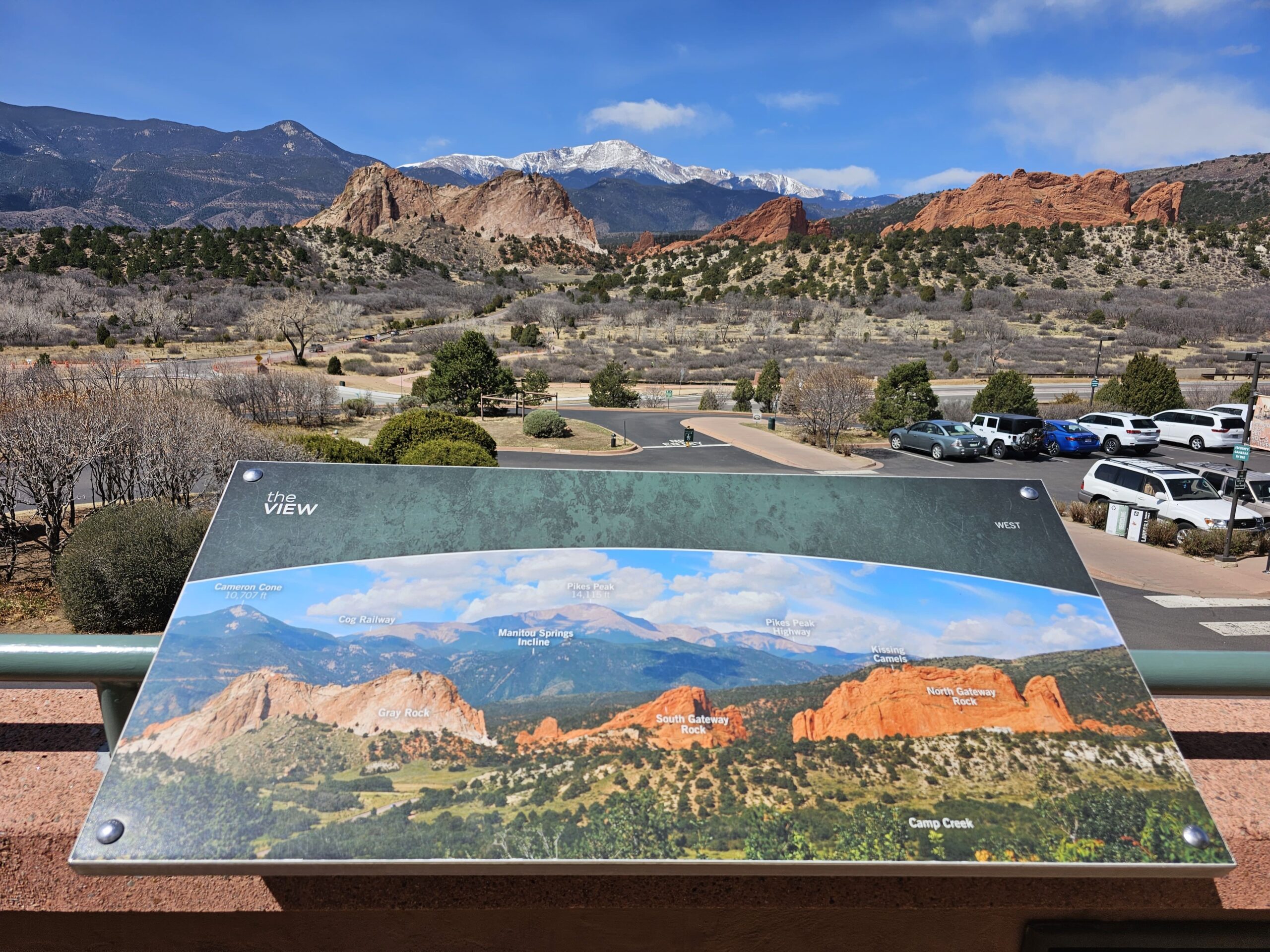 A No-Hike Way to See Garden of the Gods: Scenic Drive Tips for Older Travelers 6 Scenic overlook at Garden of the Gods Visitor Center with labeled sign pointing out landmarks like Pikes Peak, Kissing Camels, and North Gateway Rock, with snowy mountains and red rocks in the background.