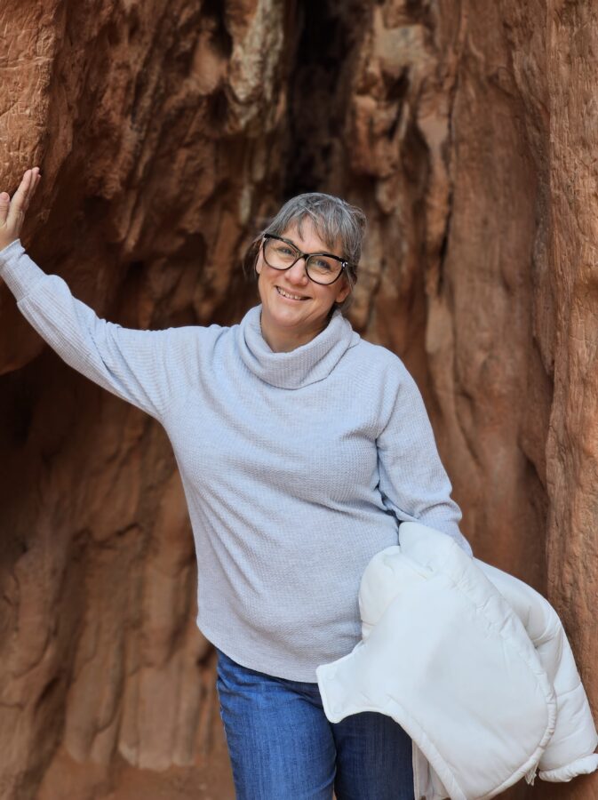 A No-Hike Way to See Garden of the Gods: Scenic Drive Tips for Older Travelers 5 Woman smiling and standing in a red rock crevice at Garden of the Gods, Colorado, wearing glasses, a light sweater, and holding a white jacket.