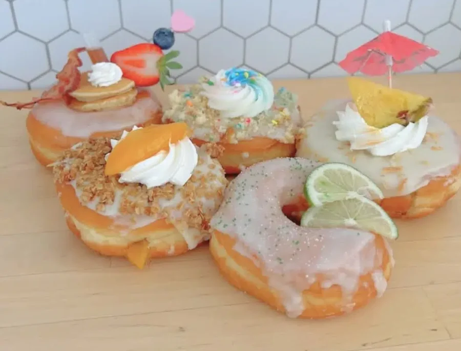 9. Fun Things to Do In Palm Coast, Florida 9 Colorful gourmet donuts topped with whipped cream, fruit slices, sprinkles, and fun decorations like a cocktail umbrella and a mini pancake, displayed on a light wood table with a white hexagon tile background.