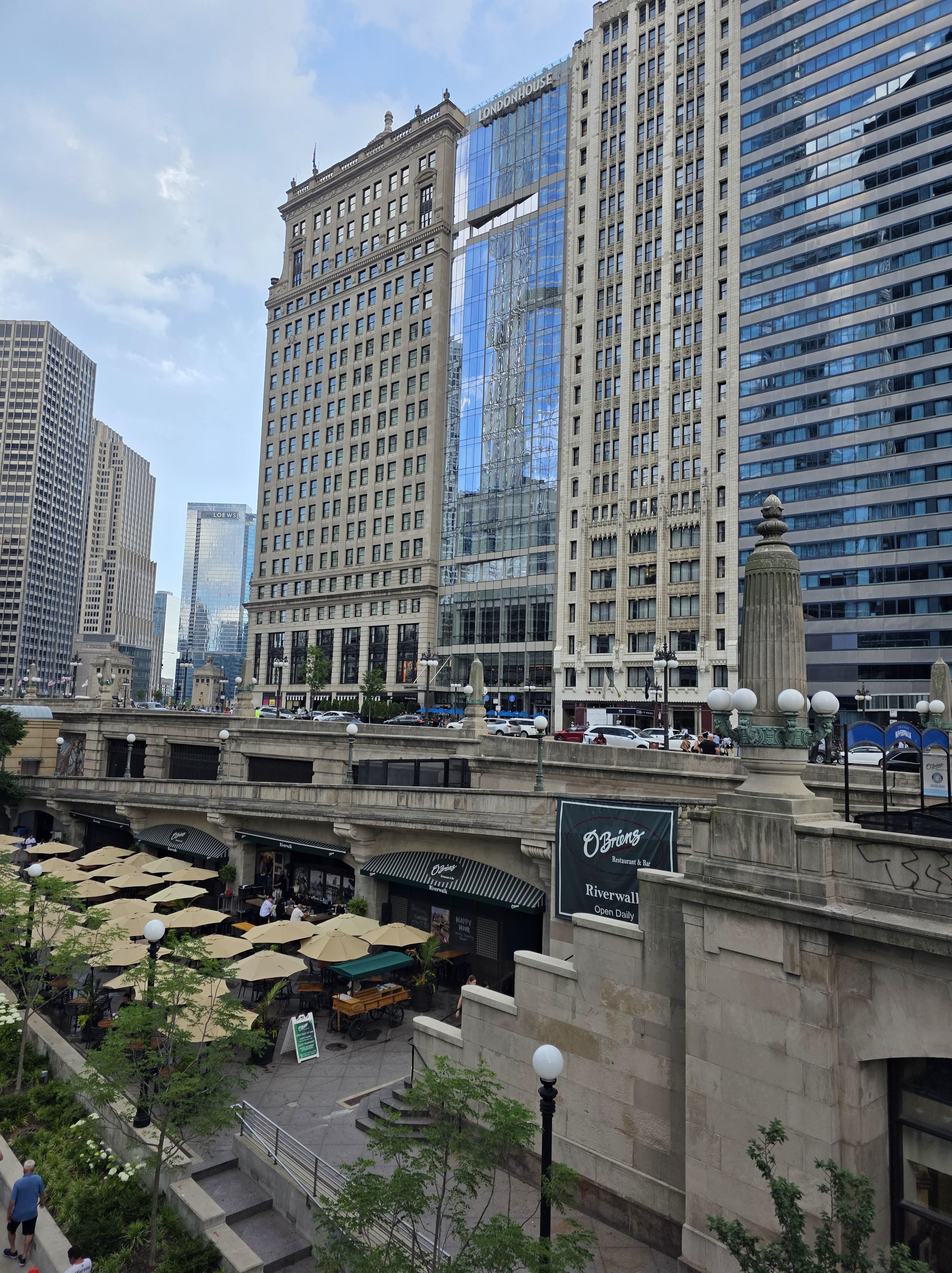 16 Best Things to Visit In Chicago 2 View of the Chicago Riverwalk lined with restaurants and umbrellas, with the historic LondonHouse Hotel in the background — a scenic spot near Navy Pier and Millennium Park.
